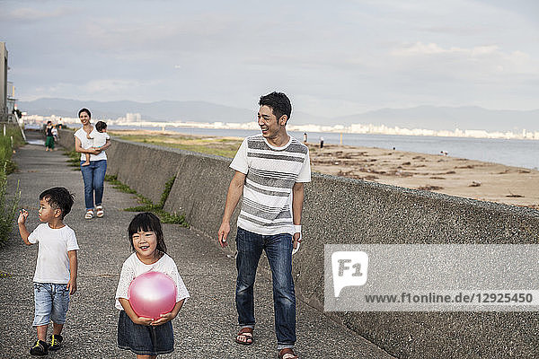 Japanese man  young boy and young girl carrying pink ball walking along concrete promenade by the ocean.