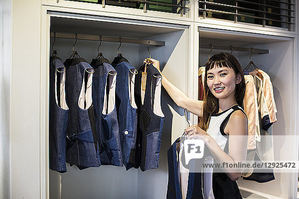 Japanische Verkäuferin steht in einem Bekleidungsgeschäft  hängt blaue Westen an eine Schiene und lächelt in die Kamera.