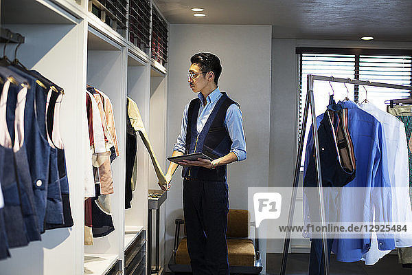 Japanese salesman with moustache wearing glasses standing in clothing store  looking at clothes on a rail  holding digital tablet.