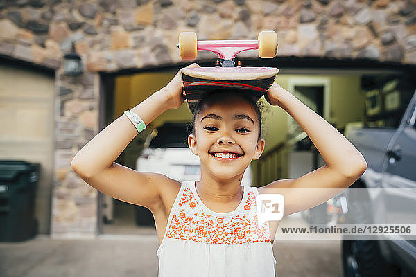 Smiling girl standing in driveway in front of house with skateboard on her head