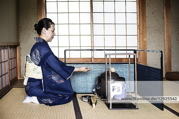 Japanerin in traditionellem hellblauem Kimono mit cremefarbenem Obi  die während einer Teezeremonie mit einem Hishaku  einer Bambusschöpfkelle  auf dem Boden kniet.