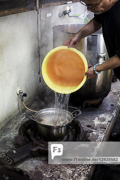 Japanese man standing in a textile plant dye workshop  pouring water from yellow plastic bucket into pot.