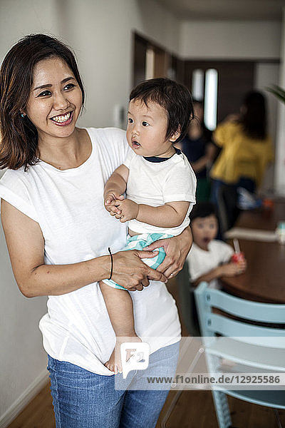 Porträt einer Japanerin  die in einem Wohnzimmer steht und ein Kleinkind trägt und in die Kamera lächelt.