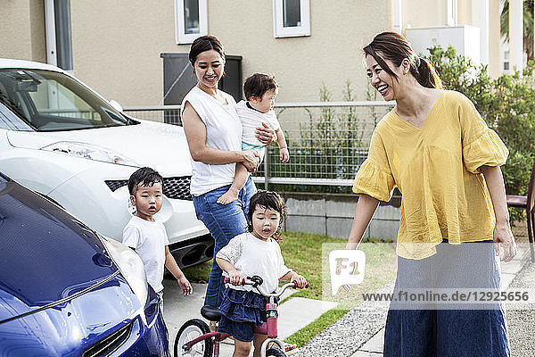 Zwei lächelnde Japanerinnen und drei kleine Kinder stehen neben einem geparkten Auto in einer Straße.