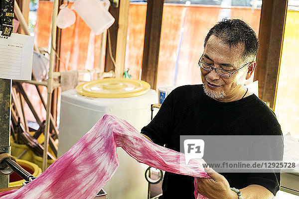 Japanischer Mann mit Brille steht in einer Textilfärberei und hält ein Stück rosa Stoff in der Hand.
