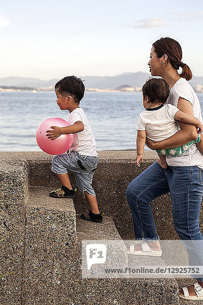 Japanese woman carrying toddler and young boy with pink ball walking up concrete steps by the ocean.