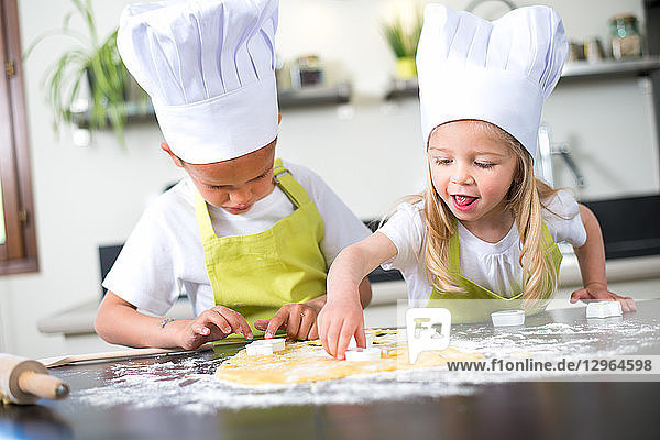 Two young kids happy childrens boy and girl family with apron and chef hat preparing funny cookies in kitchen at home.