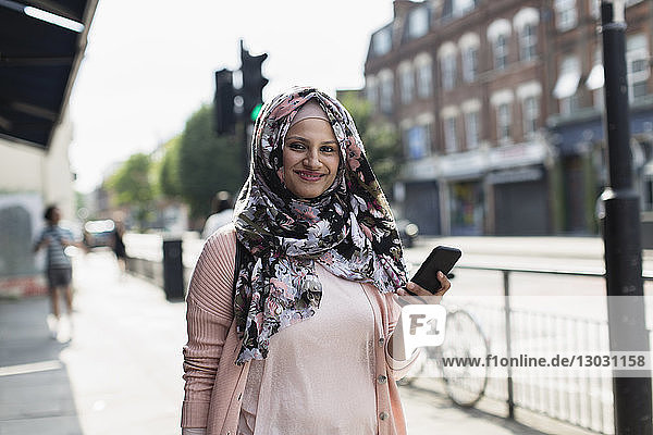 Porträt einer lächelnden  selbstbewussten Frau mit Smartphone  die einen geblümten Hijab auf einem städtischen Bürgersteig trägt