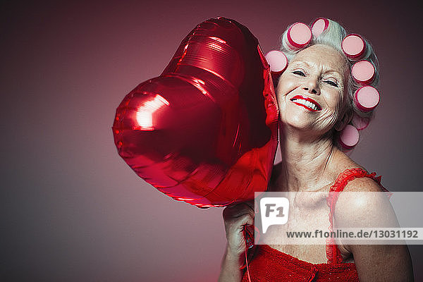 Porträt einer verspielten älteren Frau mit Lockenwicklern  die einen Luftballon in Herzform hält