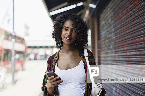 Porträt einer lächelnden  selbstbewussten jungen Frau mit Smartphone auf einem städtischen Bürgersteig