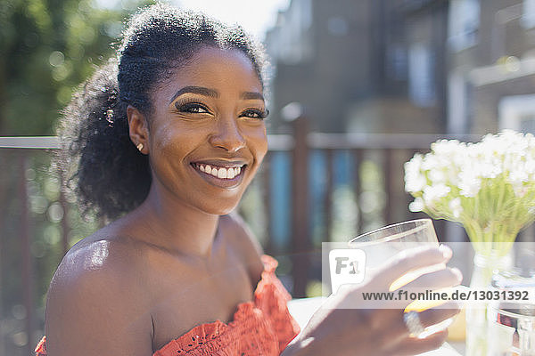 Porträt einer lächelnden  selbstbewussten jungen Frau  die auf einem sonnigen Balkon Orangensaft trinkt