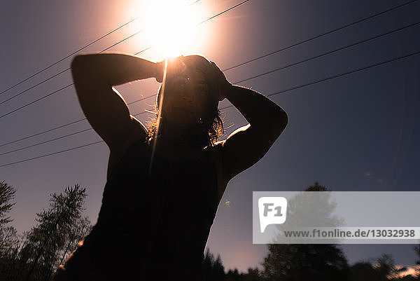 Junge Frau mit Händen auf dem Kopf gegen den sonnenbeschienenen Himmel  von hinten beleuchteter Blickwinkel