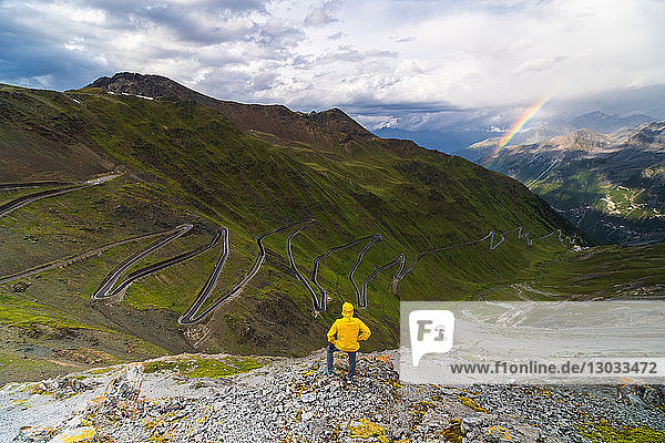 Man on rocks admires the rainbow on the winding road  Stelvio Pass  South Tyrol side  Valtellina  Lombardy  Italy