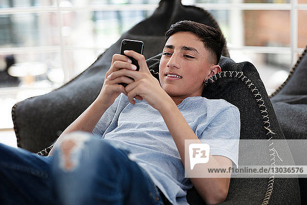 Teenage boy lying on beanbag and reading text message on cellphone