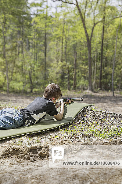 Seitenansicht eines Jungen  der im Wald auf einer Matte liegt und auf ein Gewehr zielt