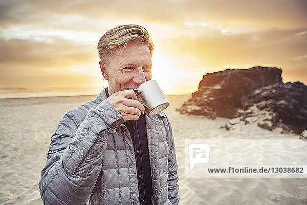 Glücklicher Mann trinkt Alkohol aus Becher  während er bei Sonnenuntergang am Strand steht