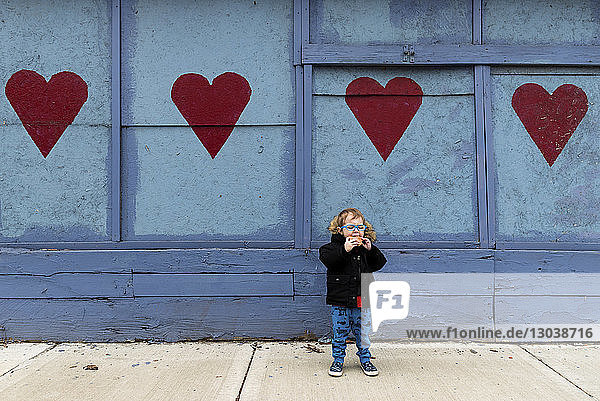Junge in voller Länge isst Nahrung  während er auf dem Fußweg gegen eine am Herzen gezogene Wand steht