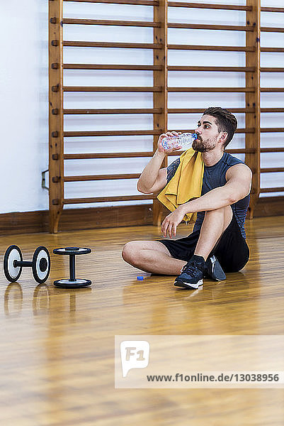 Sportler in voller Länge trinkt Wasser  während er in der Turnhalle auf einem Hartholzboden sitzt