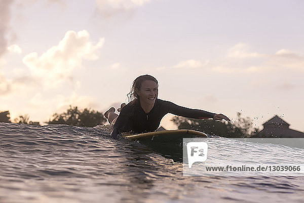Glückliche Frau liegt auf einem Surfbrett  während sie bei Sonnenuntergang auf dem Meer gegen den Himmel schwimmt