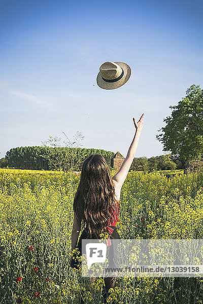 Rückansicht eines Teenager-Mädchens  das einen Hut wirft  während es am sonnigen Tag auf dem Feld vor blauem Himmel steht