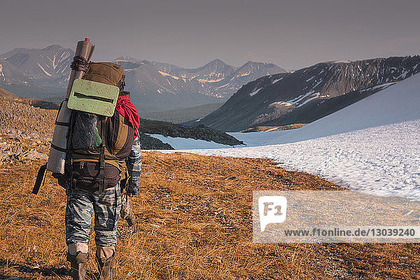 Rear view of hiker with backpack walking on field against snowcapped mountains