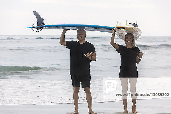 Full length portrait of smiling friends gesturing while carrying surfboards on head at beach