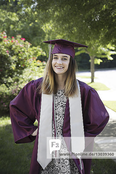 Portrait of happy woman in graduation gown standing against trees at park