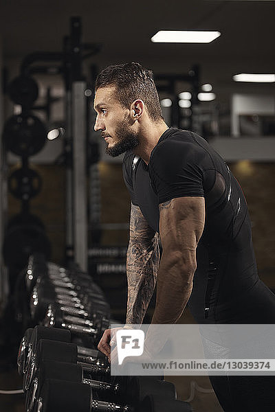 Seitenansicht eines selbstbewussten Mannes  der sich beim Training im Fitnessstudio auf Hanteln stützt