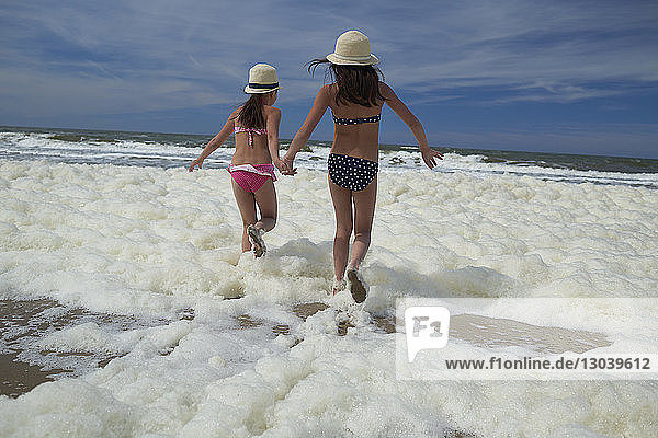 Rückansicht von Schwestern  die in Meeresschäumen laufen  während sie am Strand Händchen halten