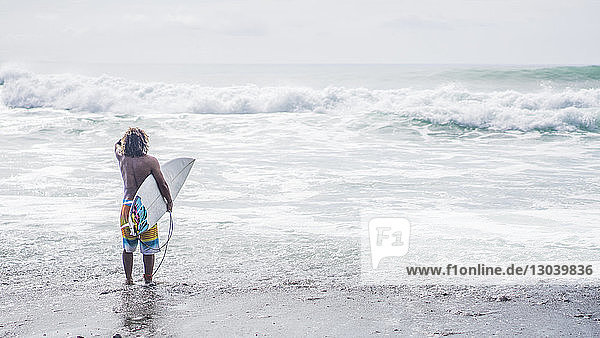 Rear view of male surfer carrying surfboard while standing on shore at beach