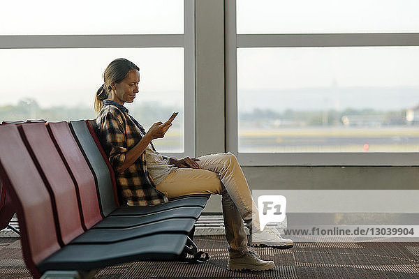 Seitenansicht einer lächelnden Frau beim Benutzen eines Mobiltelefons im Abflugbereich des Flughafens