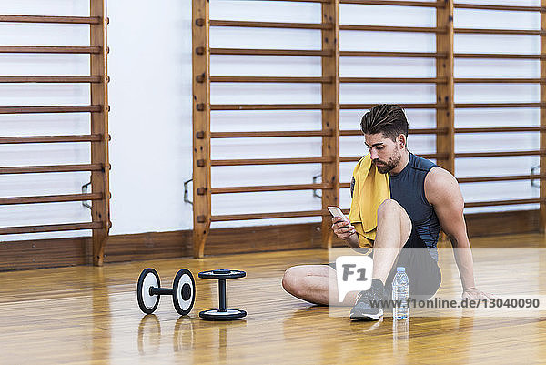 Sportler in voller Länge mit Mobiltelefon  während er mit Wasserflasche und Hanteln auf dem Hartholzboden in der Turnhalle sitzt