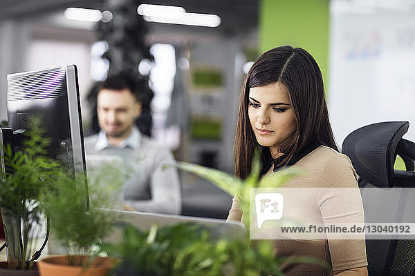 Beautiful businesswoman using laptop at desk in creative office with colleague in background