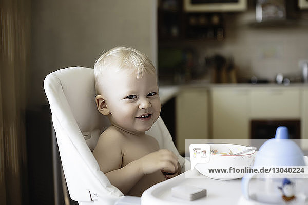 Portrait of shirtless boy having food while sitting on high chair at home
