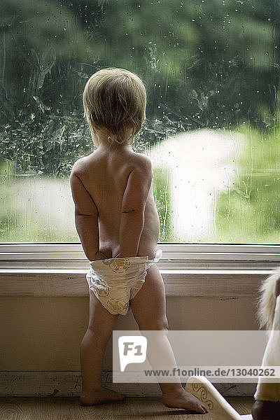 Rückansicht eines kleinen Mädchens mit Händen in Windeln  das zu Hause am Fenster steht