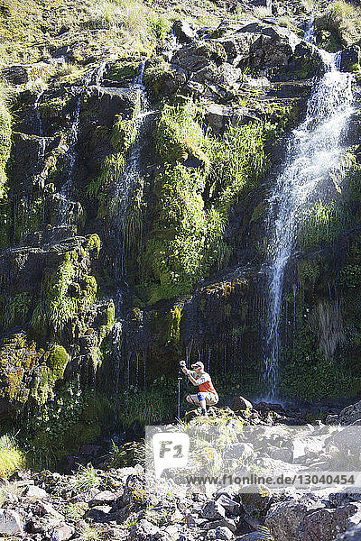 Wanderer  der bei Sonnenschein auf Felsen gegen Wasserfall und Berg kauert