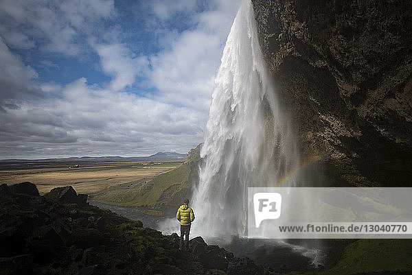 Rückansicht eines auf einem Felsen stehenden Mannes  der einen Wasserfall betrachtet