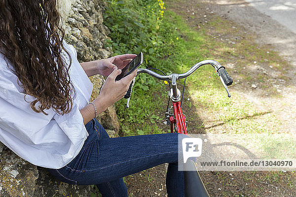 Midsection of woman using mobile phone while standing with bicycle by stone wall