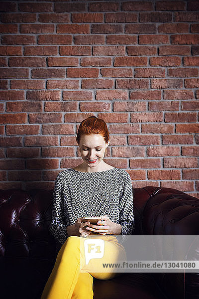 Happy businesswoman using smart phone while sitting on couch at office lobby