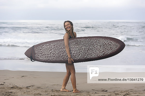 Seitenansicht einer fröhlichen Frau  die beim Strandspaziergang ein Surfbrett trägt