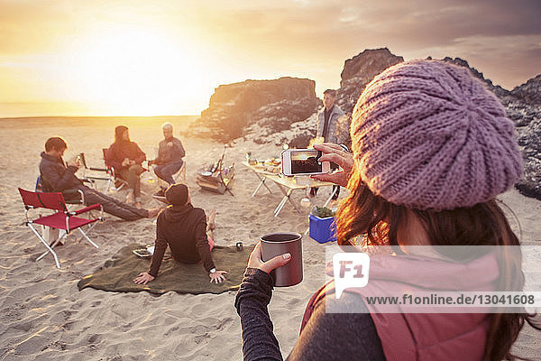 Frau fotografiert Freunde  die bei Sonnenuntergang am Strand zelten