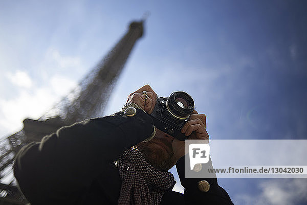 Low angle view of man photographing through DSLR camera against Eiffel Tower and blue sky
