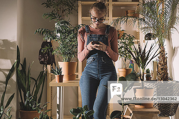 Frau benutzt Smartphone  während sie sich zu Hause an Topfpflanzen an den Tisch lehnt