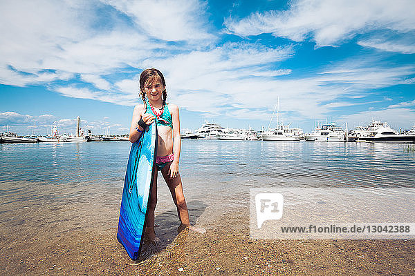 Porträt eines Mädchens  das ein Surfbrett in der Hand hält  während es am Strand am Ufer gegen den Himmel steht