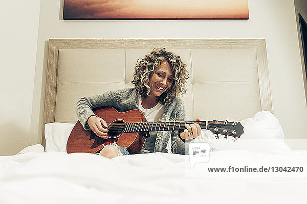 Lächelnde Geschäftsfrau spielt Gitarre  während sie im Hotelzimmer auf dem Bett sitzt