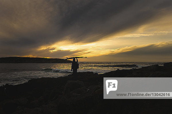 Scherenschnitt eines Mannes  der ein Surfbrett auf dem Kopf trägt  während er bei Sonnenuntergang am Strand steht