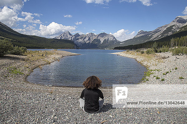 High angle view of hiker meditating while sitting at lakeshore against mountains and cloudy sky
