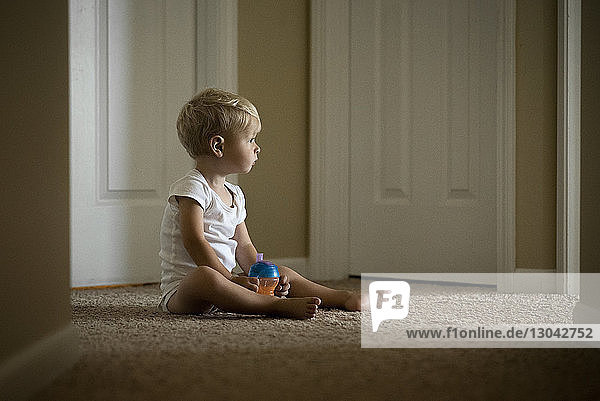Süßer Junge mit Wasserflasche schaut weg  während er zu Hause auf dem Teppich sitzt