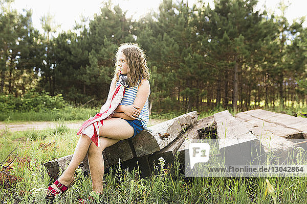 Thoughtful girl with arms crossed sitting on wood at grassy field
