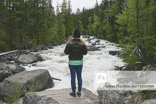 Rückansicht einer Frau in warmer Kleidung  die auf einem Felsen am fließenden Fluss im Wald steht